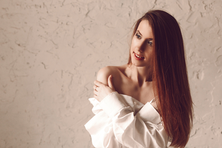 Portrait of attractive young redhead woman with long hair sitting in man's shirt touching herself for naked shoulderの写真素材
