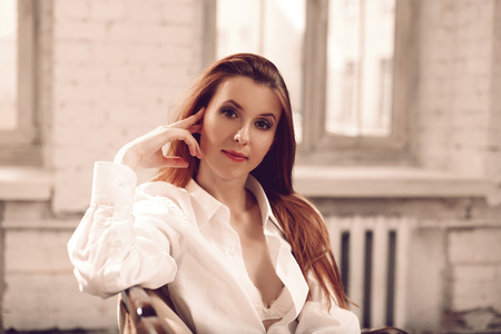 Beautiful young woman with long hair sitting on sofa in bra under unbuttoned man's shirtの写真素材