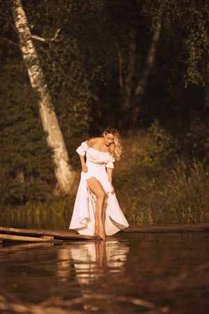 Beautiful young woman with gorgeous long blond hair standing on wooden platform by the lake at sunset or sunrise showing her sexy legsの写真素材