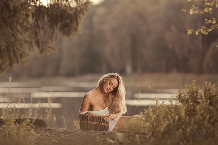 Lovely young woman with beautiful breasts sitting by the lake at sunset and holding picnic basketの写真素材