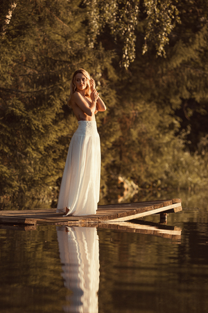 Attractive young woman with beautiful nude body standing on wooden platform by the lake at sunset or sunrise covering breasts by her handsの写真素材