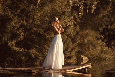 Attractive young woman with beautiful nude body standing on wooden platform by the lake at sunset or sunrise covering breasts by her handsの写真素材