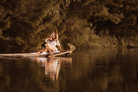 Cute young woman sitting on wooden platform by the lake at sunset or sunrise touching water by her legの写真素材