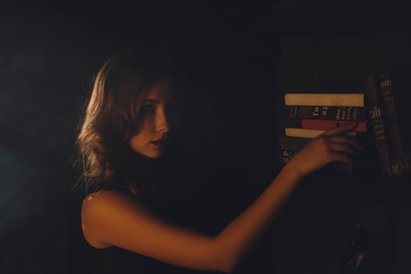 Beautiful young woman standing in a dark room pointing on a book on a bookshelfの写真素材