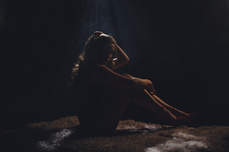Silhouette of beautiful young woman with a long hairs sitting leaning on her knees and looking in camera on a darck background.の写真素材