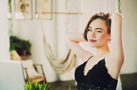 Attractive young woman in black lace lingerie body sitting and relaxing at sunny morning, working on notebook or laptop onlineの写真素材