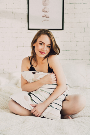 Attractive young woman in sexy black lingerie body sitting in white bed hagging a pillow and flirting with a cameraの写真素材