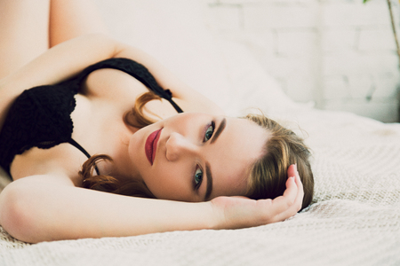 Sensual young woman with red sexy lips lying in white bed in black lace lingerie body and looking in cameraの写真素材