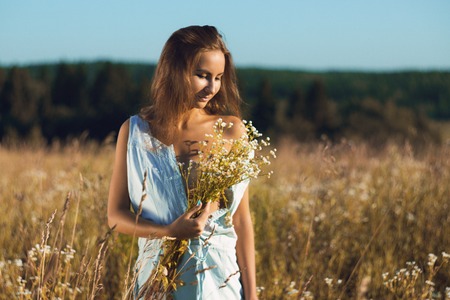 Sensual young girl in sleeveless jean sundress standing in field of tall grass covering naked breasts by bouquetの写真素材