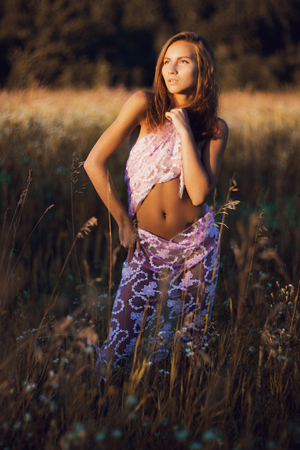 Attractive young woman with naked slim body covered with silk scarf standing in a summer meadow enjoing sunsetの写真素材