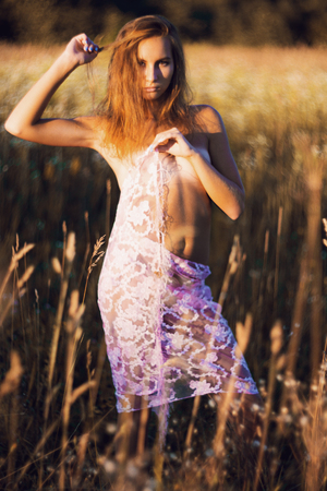 Attractive young woman with naked slim body standing posing in a summer meadow on sunset covering her small breasts and hips with silk scarfの写真素材