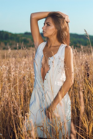 Sensual young girl with slim body standing and posing in the meadow in unbuttoned sleeveless jean sundressの写真素材