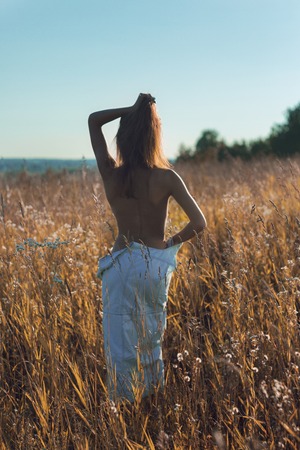 Pretty young woman in sleeveless jean sundress with naked back standing alone in a beautiful summer field enjoying the sunの写真素材
