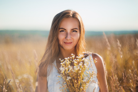 Attractive young woman standing smiling in the meadow in sleeveless jean sundress looking in cameraの写真素材