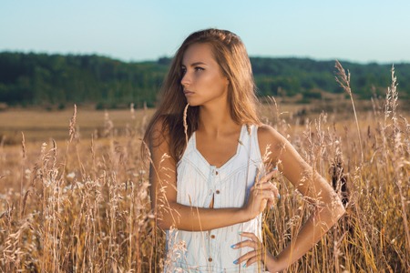 Sensual young girl in sleeveless jean sundress standing posing in the field of tall grass on sunsetの写真素材