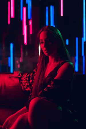 Attractive young woman sitting on a leather sofa lit by red neon light and with a wall of neon tubes in a backgroundの写真素材