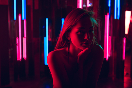 Portrait of beautiful young woman sitting leaning on her hand against a wall of neon lightsの写真素材