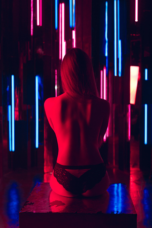 Sexy young woman in sexy lingerie sitting on mirror cube with naked back against a wall of neon tubesの写真素材