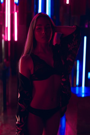 Attractive young woman standing in sexy teasing pose in black lingerie against a wall of neon lightsの写真素材