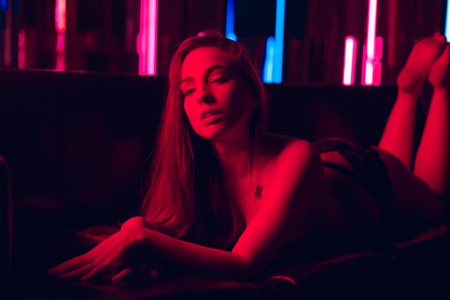 Beautiful young woman lit by red neon light sexy bending over lying naked or nude on a sofa against a wall of neon tubesの写真素材