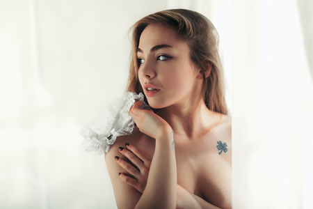Topless young woman with a garter in hands in white curtainsの写真素材