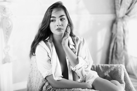 Attractive young woman with long hair is sitting in chair topless in unbuttoned man's shirtの写真素材
