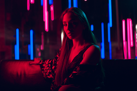 Sensual young woman sitting on a leather sofa lit by red neon light and with a wall of neon tubes in a backgroundの写真素材