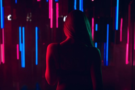 Silhouette of beautiful young woman staying topless against a wall of neon light showing her beautiful naked backの写真素材