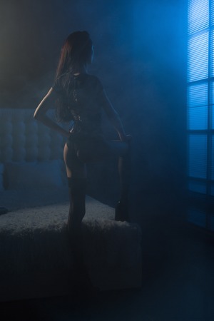 Attractive young woman in a sexy witch costume and torn stockings getting ready to halloween party in her bedroom in front of window with moonlightの写真素材