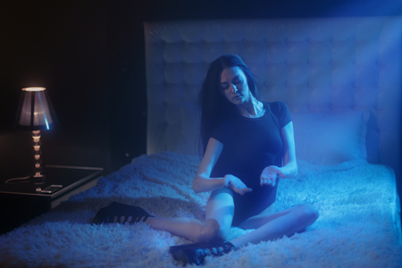 Sexy young woman with black hair and naked long legs sitting on white bed and playing with a magick moon light falling onto her hands, wearing sexy black lingerie as a witch costume before Hellowing party.の写真素材