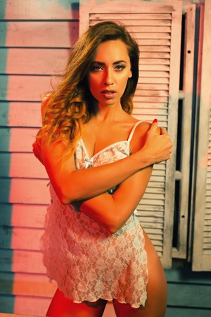 Attractive young woman with sensual athletic body standing hugging herself in transparent lace chemise at beach house near ocean side at sunsetの写真素材