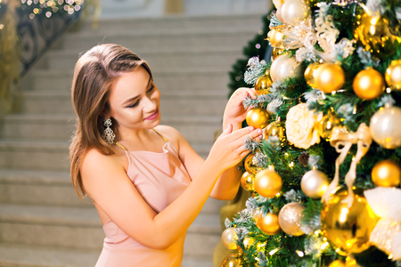 Beautiful young woman in a pink elegant evening dress staying in a chic hall and dressing up a christmas tree with yellow ball on a new year eve.の写真素材