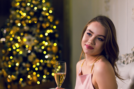 Cute young woman is sitting in a dark chic hall with a Christmas tree dressed up in a garland in ampir chair with a glass of champagne in her hands enjoying new year holidays.の写真素材