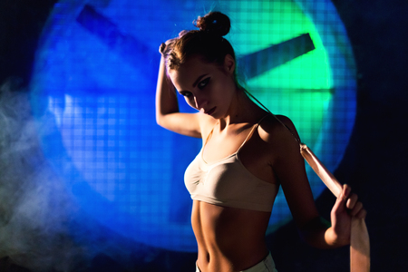 Silhouette of beautiful young gymnast woman with a slim sport body posing with gymnastics tape on a dark background against the black wall with a big fan lit with blue and green neon light.の写真素材
