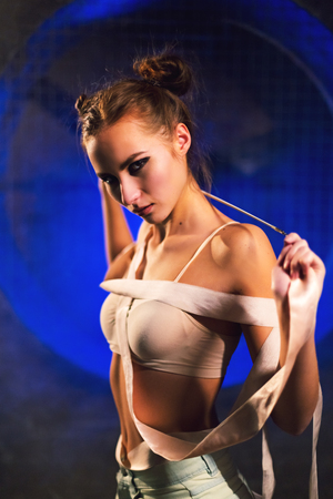 Sultry beautiful young gymnast woman with a slim sport body posing with gymnastics tape on a dark background against the black wall with a big fan lit with blue neon light.の写真素材