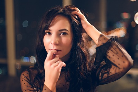Portrait of a beautiful young woman in black cocktail dress leaning on a wall in hotel room with owesome view on night city from a window and served dining table.の写真素材
