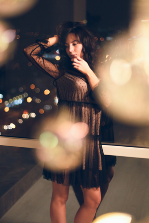 Sensual young woman in black lace cocktail dress standing in alluring pose in front of window with owesome view on a night city.の写真素材