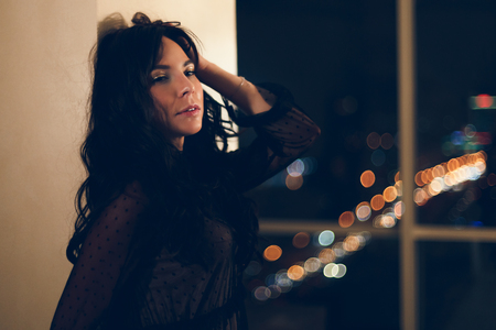 Portrait of a attractive young woman in black lace cocktail dress leaning on a wall in hotel room with owesome view on night city from a window and served dining table.の写真素材