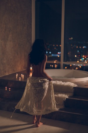 Sensual young woman staying naked or nude with white lace robe on her hips getting ready to take a bath in luxury hotel room at night with awesome city lights view trough big window.の写真素材