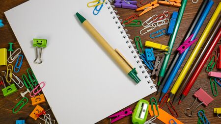 Paper notebook with green pen. School stationery on brown wooden table: multi-colored pencils, paper...