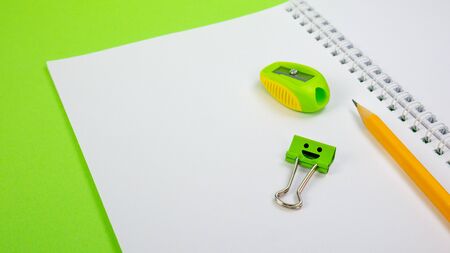 School Notepad with Yellow Pencil, Pencil Sharpener, Smile Binder Clip on Green Background. Spiral...
