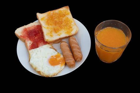 Breakfast on the table consists of toast, topped with orange jam and strawberry jam, sausage, grilled fried egg and squeezed orange juice.の写真素材
