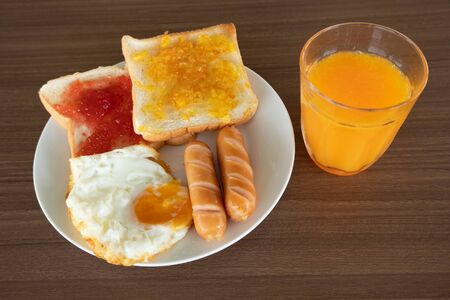 Breakfast on the table consists of toast, topped with orange jam and strawberry jam, sausage, grilled fried egg and squeezed orange juice.の写真素材