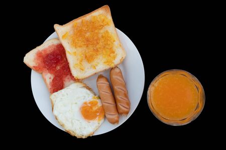 Breakfast on the table consists of toast, topped with orange jam and strawberry jam, sausage, grilled fried egg and squeezed orange juice.の写真素材