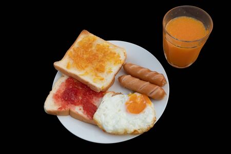 Breakfast on the table consists of toast, topped with orange jam and strawberry jam, sausage, grilled fried egg and squeezed orange juice.の写真素材