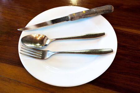 Empty white plate with knife and fork, spoon, on wooden table.の写真素材