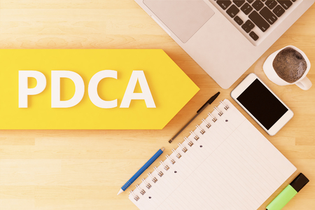 PDCA - Plan Do Check Act - linear text arrow concept with notebook, smartphone, pens and coffee mug on desktop - 3d render illustration.の写真素材