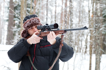 Hunter in a fur cap with ear flaps with sniper rifle in winter forest.