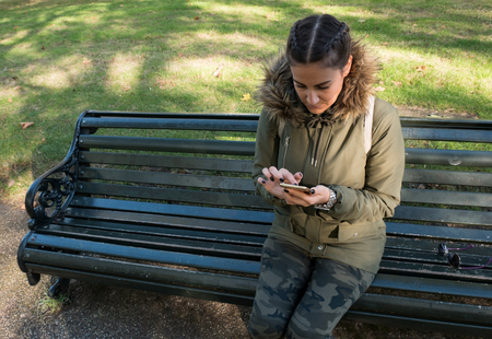 Young woman typing message on smart phone on bench in the parkの写真素材