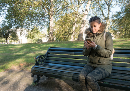 Young woman sitting on the bench in park and typing message on smart phoneの写真素材